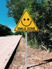 Semn de circulație galben cu o față zâmpitoare pe care scrie în limba engleză:„Loc de zâmbit”